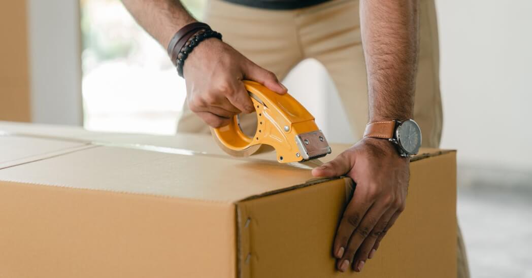 Crop faceless young male with wristwatch using adhesive tape while preparing cardboard box for transportation