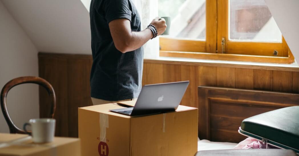 Side view of young ethnic male owner of newly bought house with cup of coffee in bedroom full of boxes and suitcases standing and looking at neighboring houses through window