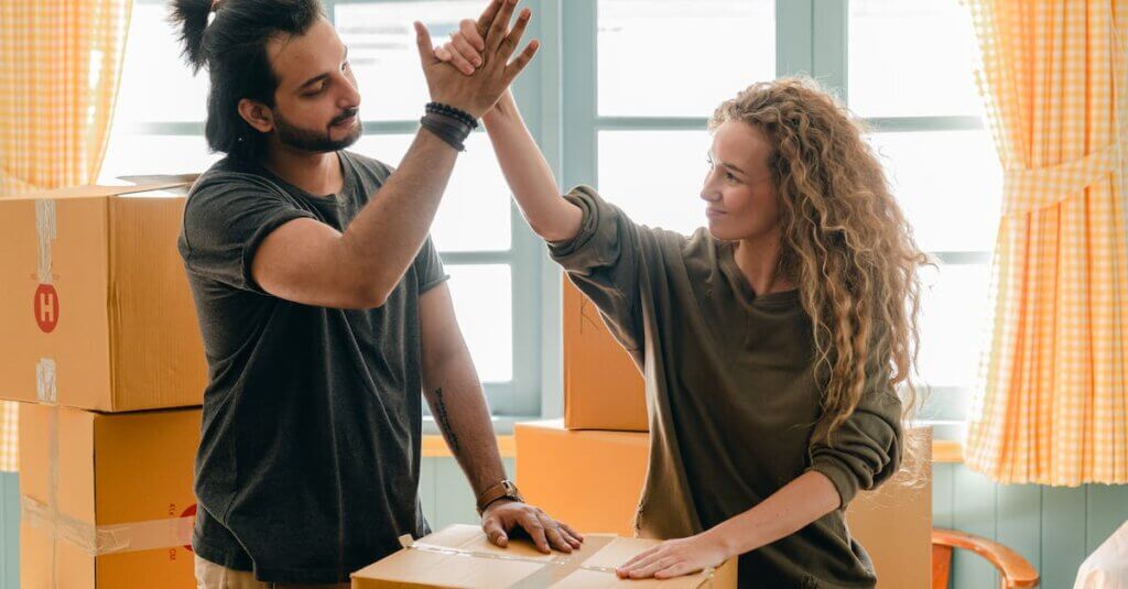 Happy woman in casual wear standing near heap of cardboard boxes and giving high five to ethnic boyfriend with ponytail showing agreement while looking at each other